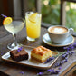 A brownie, lemon bar, focaccia bread, martini with a lavender sprig, lemonade, steaming cup of latte all on a rustic wooden table with lavender sprigs laying around on it